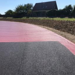 ADAMS - Nowo wybudowana droga z nawierzchnią asfaltową w kolorze czarnym i czerwonym, otoczona zielenią i brązową skarpą, w tle dom jednorodzinny.