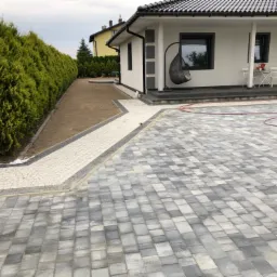 Nowoczesny podjazd z szarej kostki brukowej prowadzący do domu z białymi ścianami i ciemną dachówką, obok ścieżka z jasnej kostki i rząd tuj.