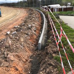 ADAMS - Wykop pod krawężnik na placu budowy drogi, widoczne przygotowane podłoże, betonowe obrzeża i taśmy ostrzegawcze, w tle budynki gospodarcze i ogrodzenie.