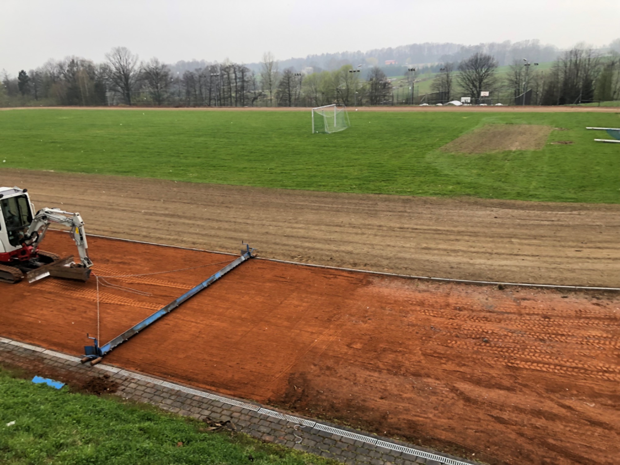 Mini koparka ciągnie za sobą specjalistyczny sprzęt do wyrównywania nawierzchni kortu tenisowego z ceglanej mączki na tle boiska sportowego.