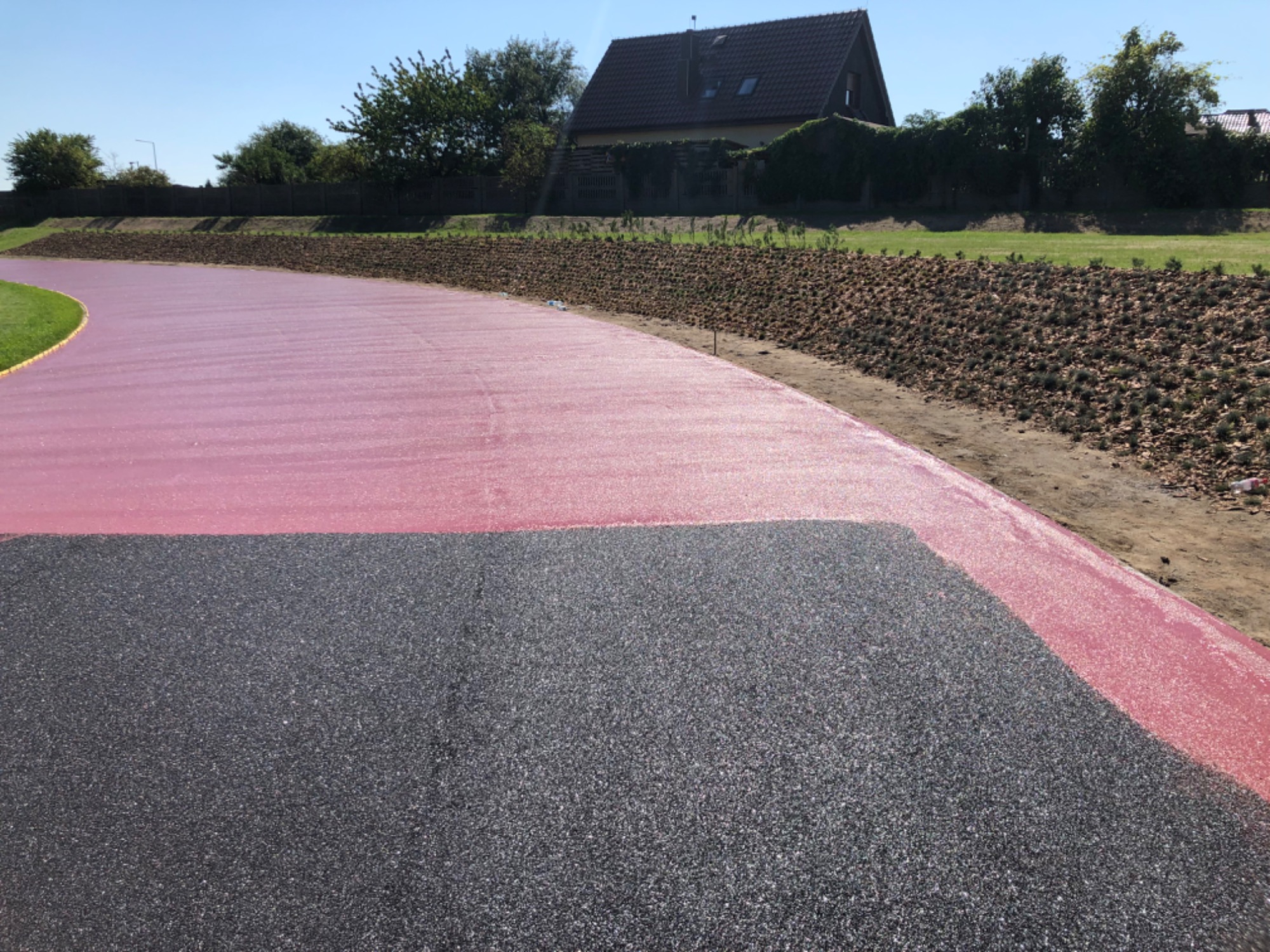 Nowo wybudowana droga z nawierzchnią asfaltową w kolorze czarnym i czerwonym, otoczona zielenią i brązową skarpą, w tle dom jednorodzinny.