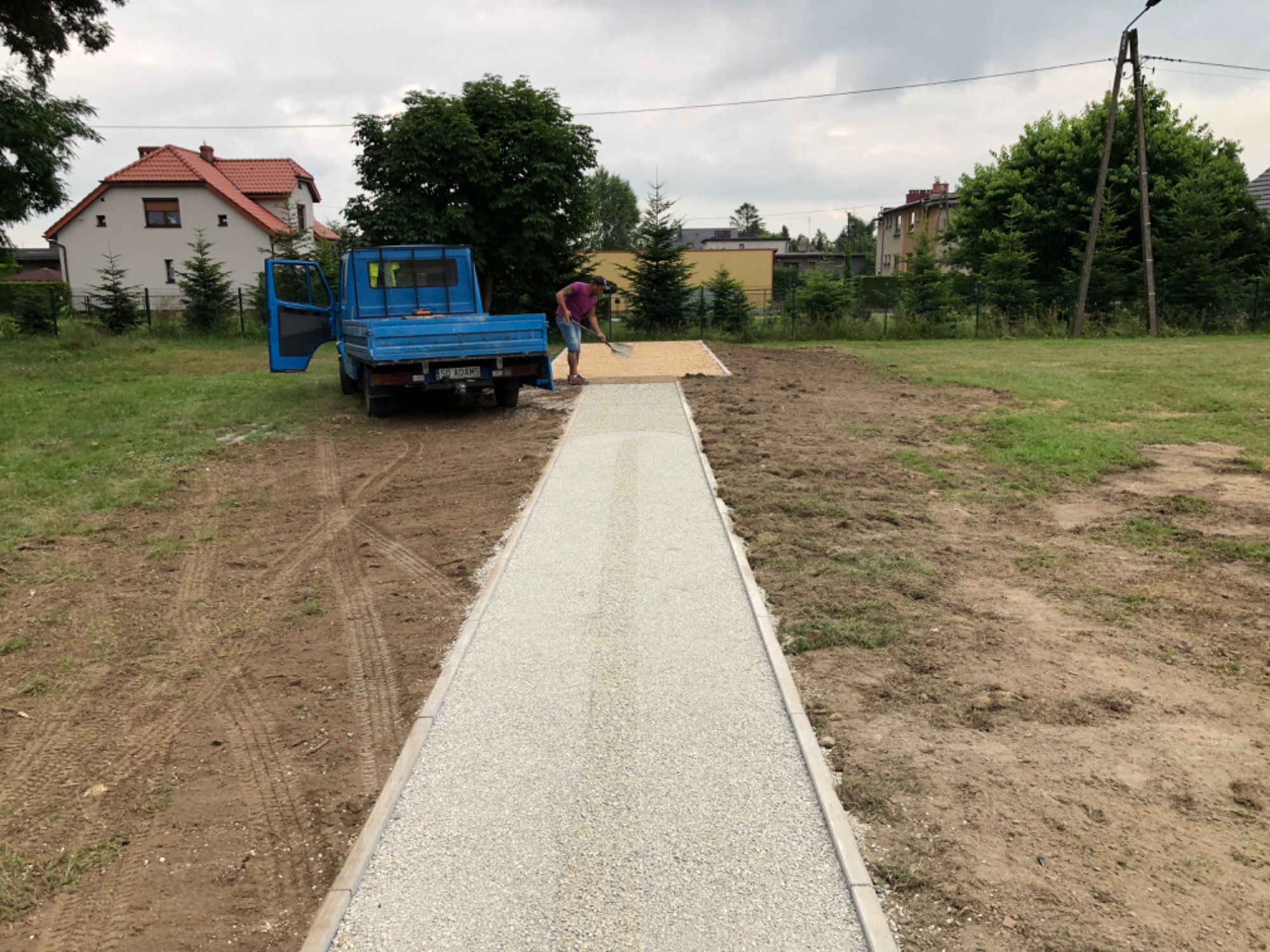 Pracownik wyrównuje szutrową nawierzchnię drogi szpachlą, niebieska ciężarówka z otwartymi drzwiami stoi obok, w tle zabudowania mieszkalne i zieleń