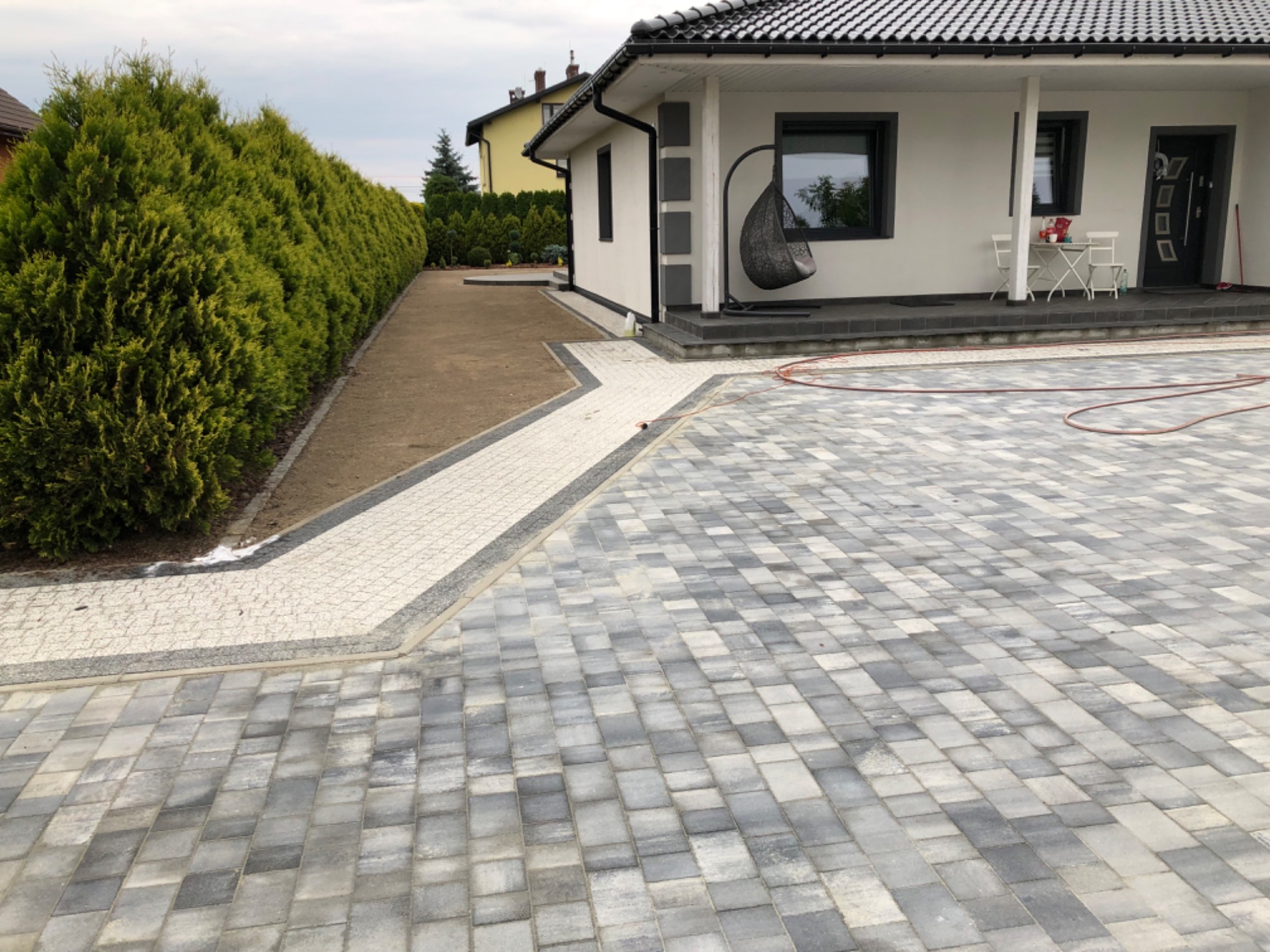 Nowoczesny podjazd z szarej kostki brukowej prowadzący do domu z białymi ścianami i ciemną dachówką, obok ścieżka z jasnej kostki i rząd tuj.