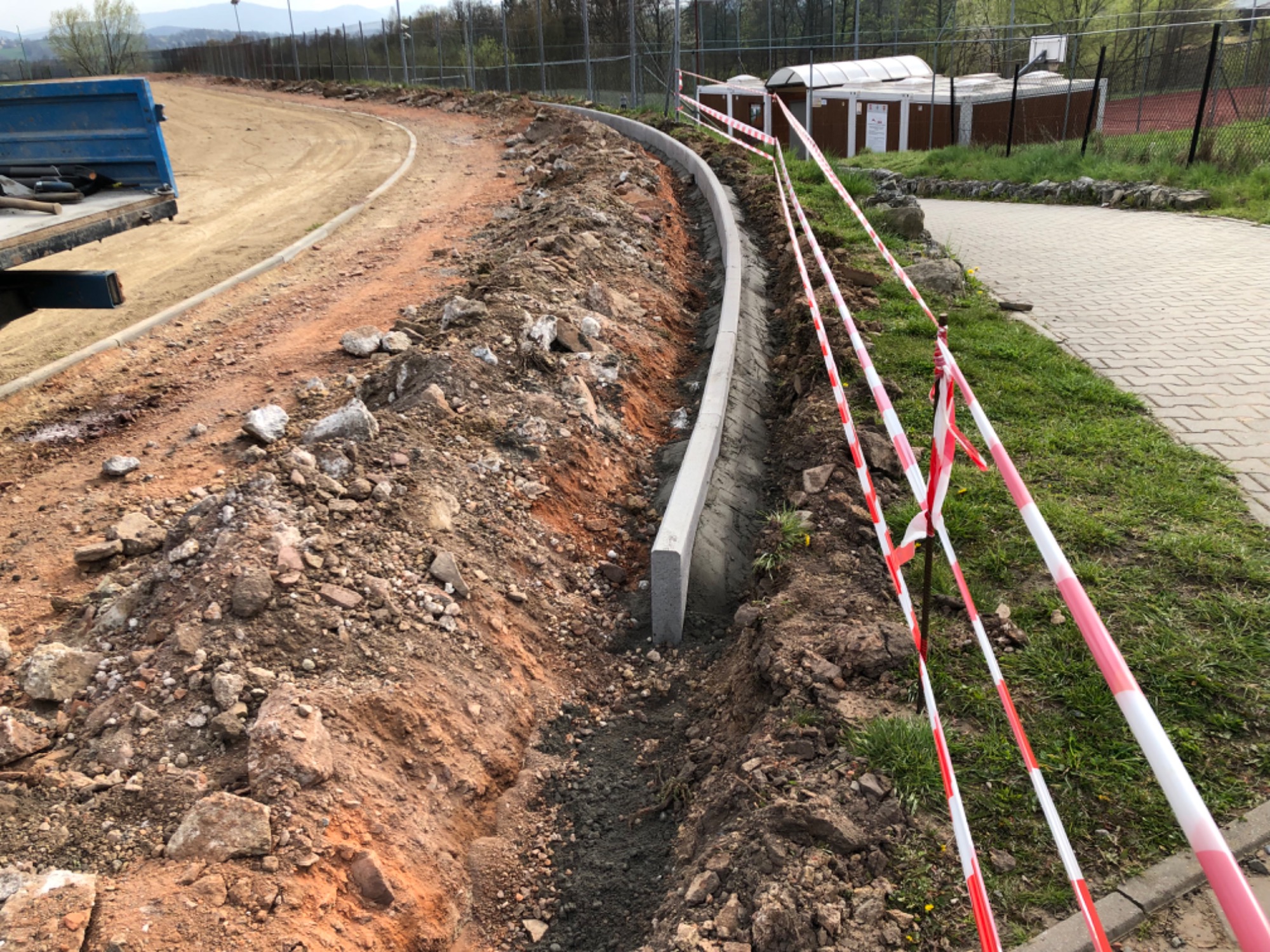 Wykop pod krawężnik na placu budowy drogi, widoczne przygotowane podłoże, betonowe obrzeża i taśmy ostrzegawcze, w tle budynki gospodarcze i ogrodzenie.