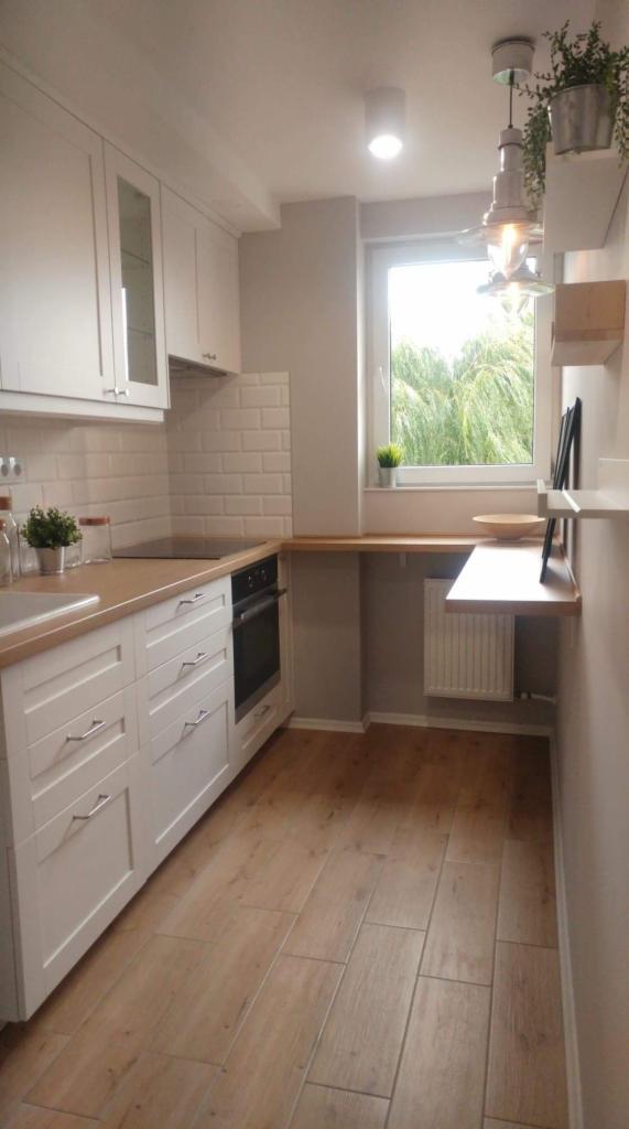 Jasna kuchnia w Warszawie z białymi szafkami, drewnianym blatem i panelami podłogowymi imitującymi drewno, wykończona białymi płytkami typu metro, z oknem wychodzącym na zieleń.