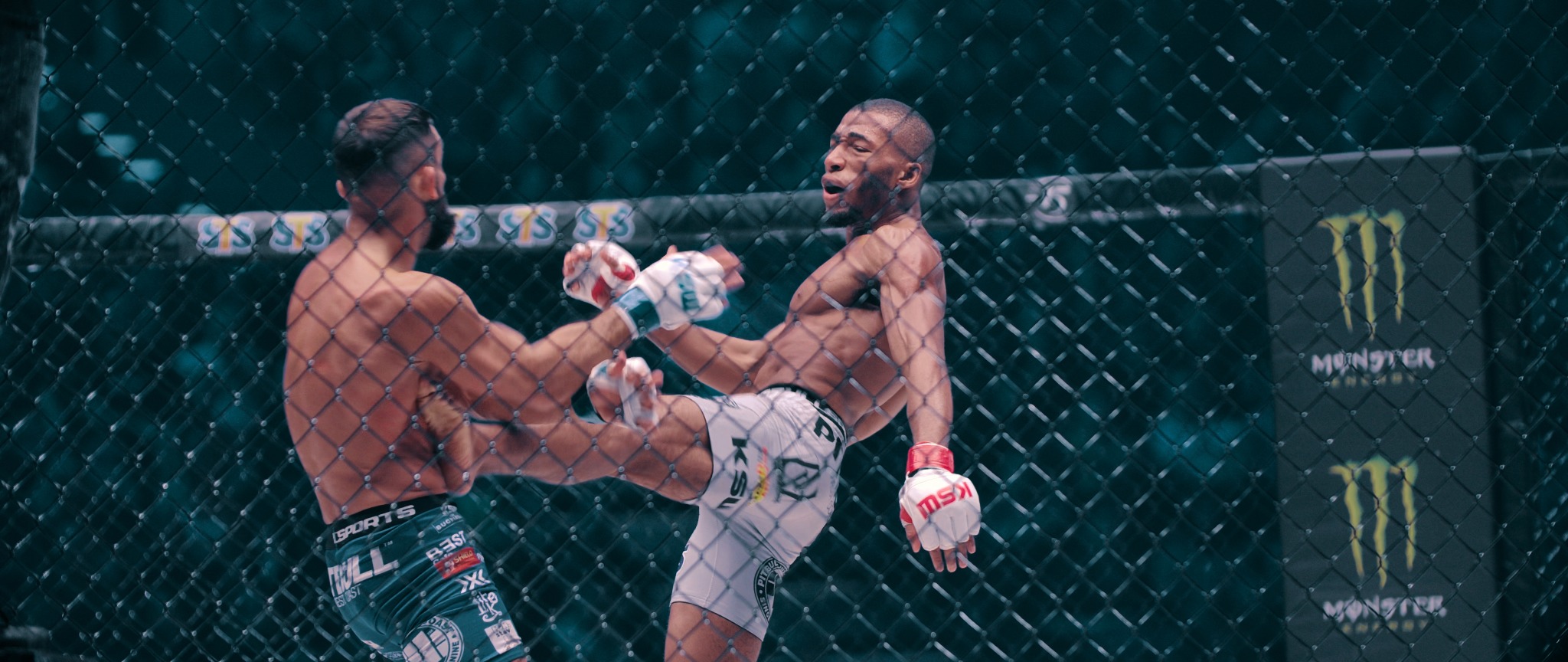 Dwóch zawodników MMA w ringu podczas walki, jeden z nich wykonuje kopnięcie, widoczne logo Monster Energy w tle.