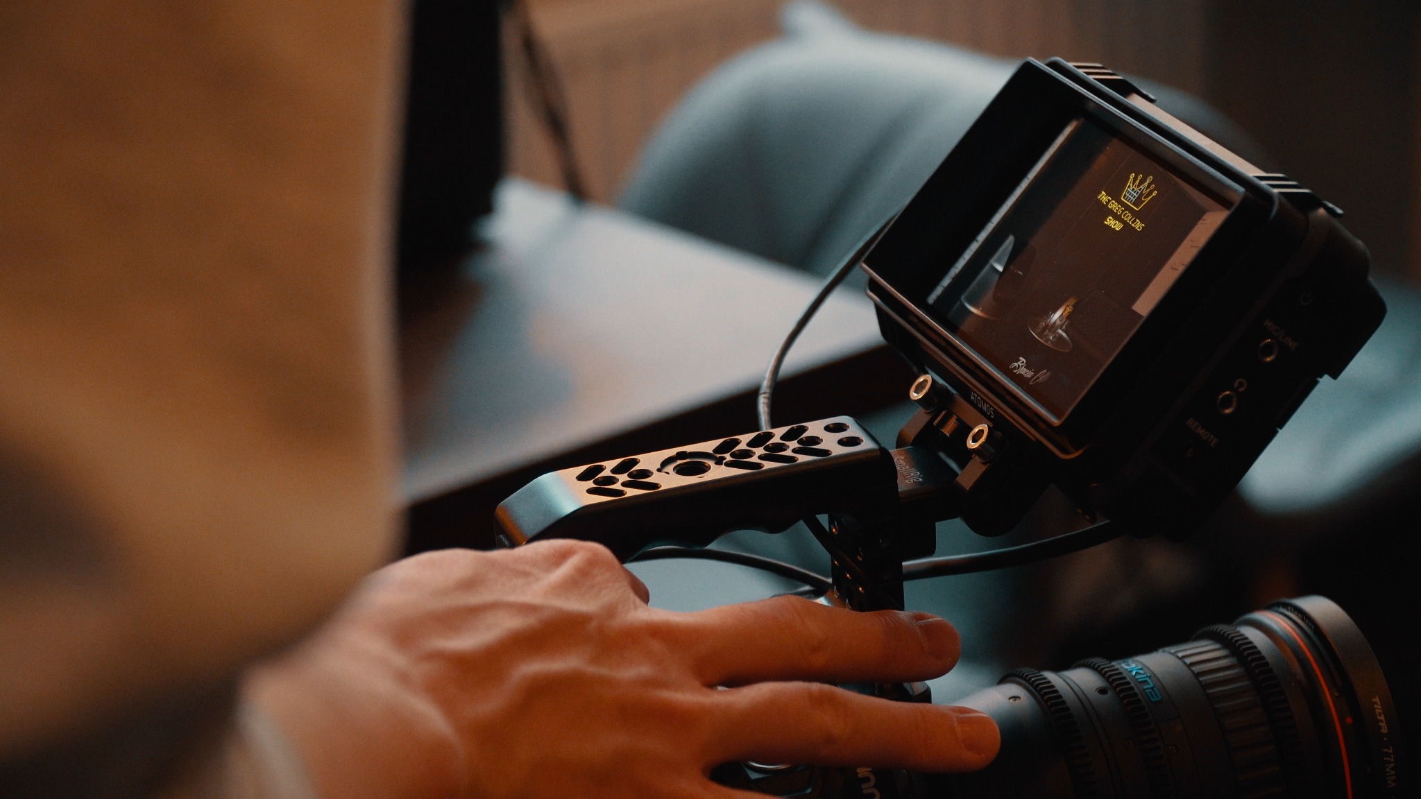 Operator filmowy trzymający profesjonalną kamerę z monitorem Atomos, wyświetlającym logo 'The Greg Collins Show', w tle rozmyte wnętrze.