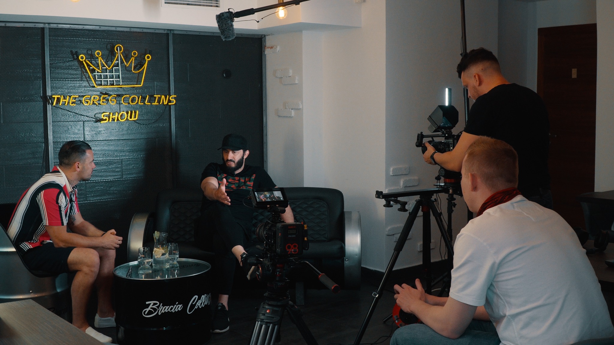 Ekipa filmowa kręci wywiad w studio z neonowym logo 'The Greg Collins Show' w tle. Operator kamery z oświetleniem i mikrofon kierunkowy nad głową osoby przeprowadzającej wywiad.