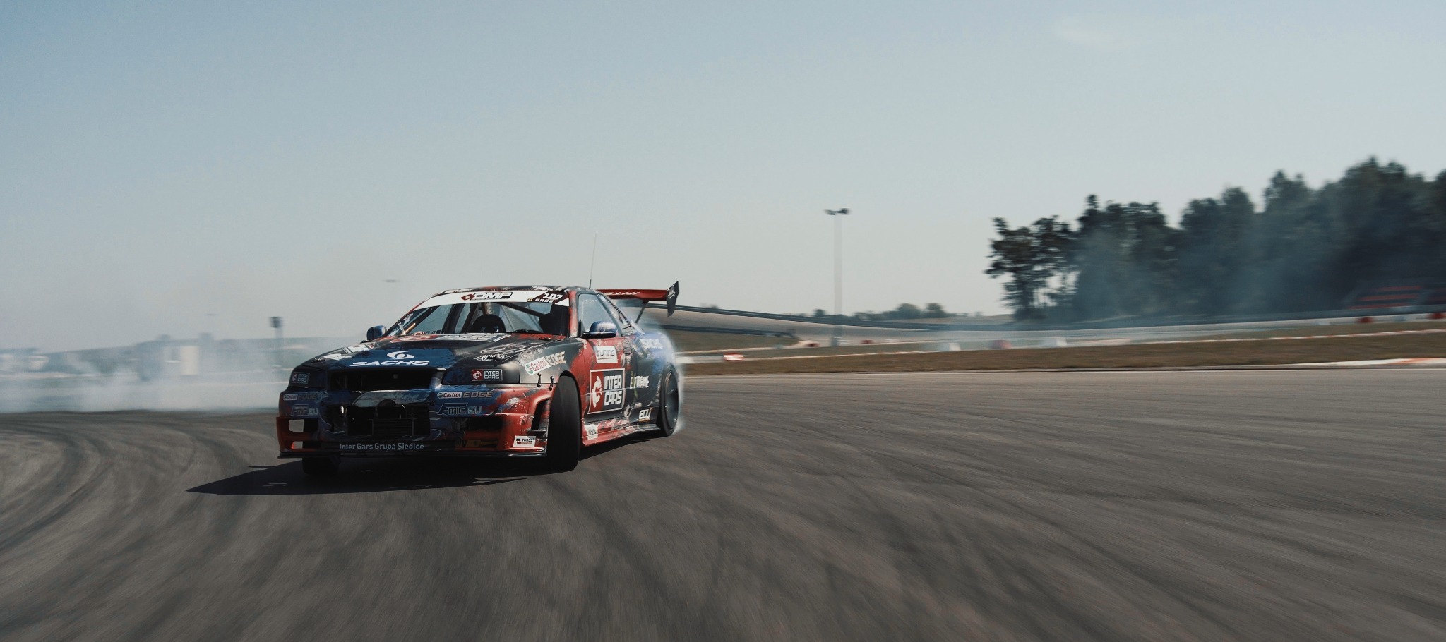 Driftingowy samochód z naklejkami sponsorów, pędzący po torze wyścigowym, z dymem unoszącym się z opon, w tle zalesiony teren.