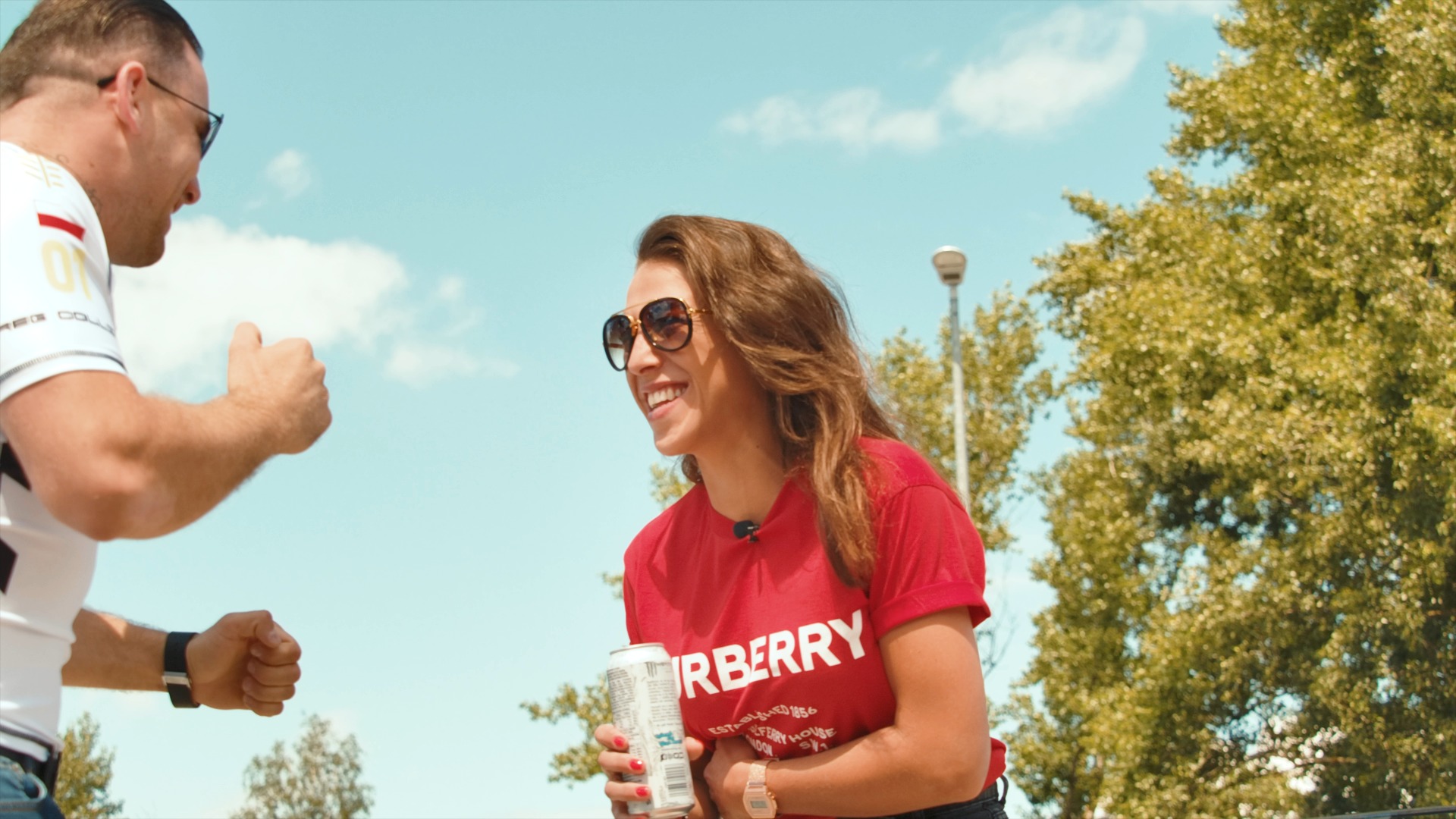 Uśmiechnięta kobieta w czerwonej koszulce z napisem trzymająca puszkę napoju, rozmawiająca z mężczyzną w okularach, który unosi kciuk w górę, na tle błękitnego nieba i drzew.