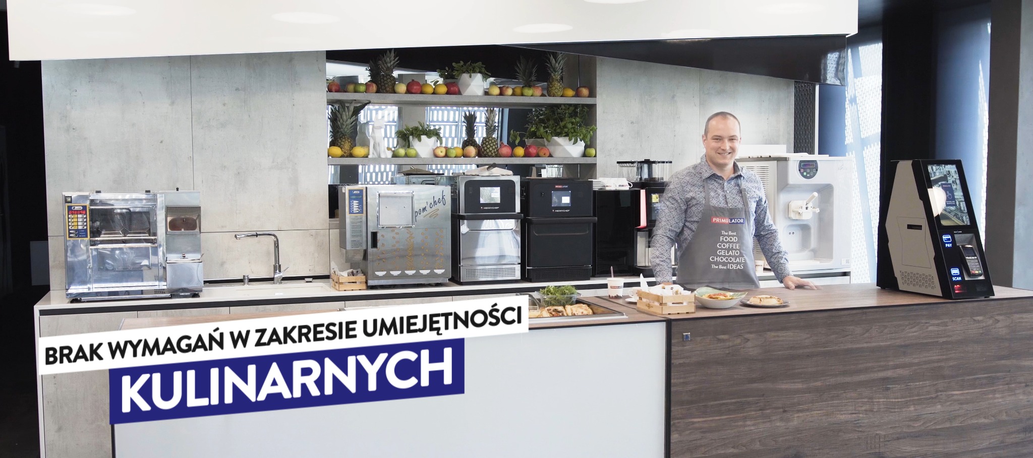 Uśmiechnięty mężczyzna w fartuchu z napisem 'PRIMULATOR' stoi za blatem z ekspresami do kawy i lodów, na ścianie półka z owocami.