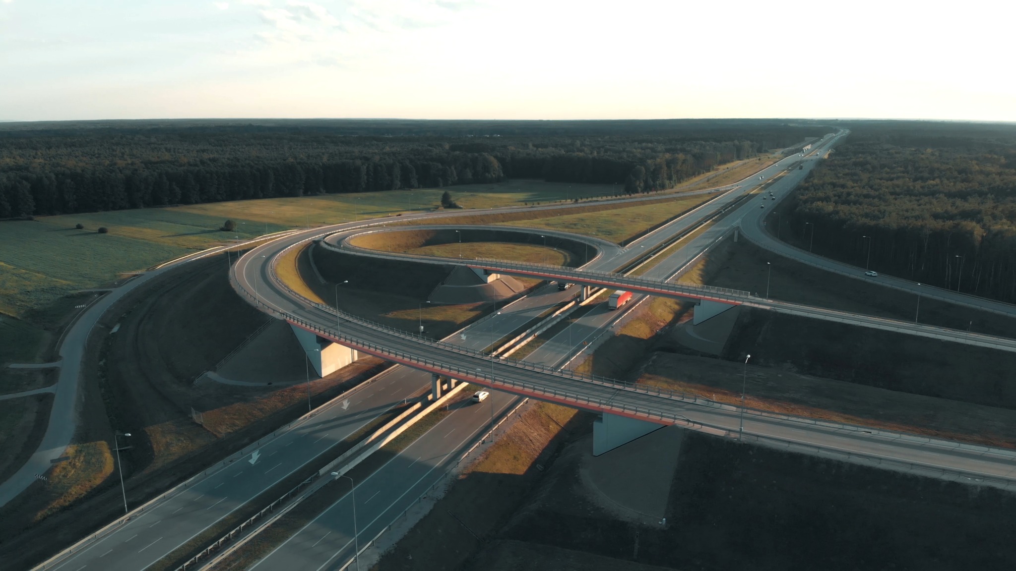 Widok z lotu ptaka na skrzyżowanie autostrad z wieloma wiaduktami, otoczone zielenią i lasami, oświetlone popołudniowym słońcem, z widocznymi samochodami i ciężarówką.