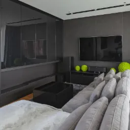 Nowoczesny salon w odcieniach szarości z zabudową ściany z czarnymi, lakierowanymi frontami, szarą sofą z licznymi poduszkami i dekoracyjnymi kulami z mchu.