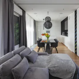 Nowoczesne wnętrze: szary narożnik z poduszkami i pledem, stół z czarnymi krzesłami, kuchnia z białymi frontami w tle, drewniana podłoga, designerskie lampy wiszące i logo firmy w prawym dolnym rogu.