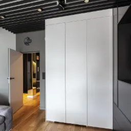 Nowoczesna szafa wnękowa z białymi frontami, wkomponowana w aranżację salonu z drewnianą podłogą i designerskim oświetleniem sufitowym.