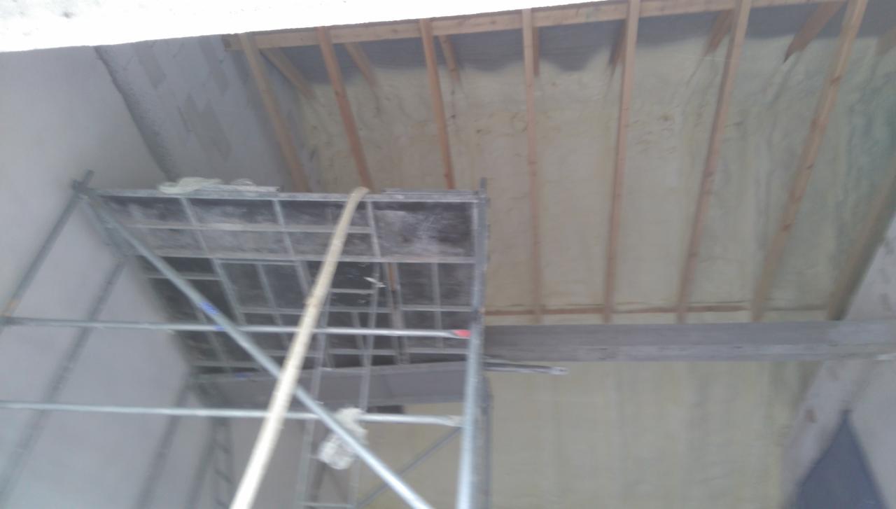 Poddasze z drewnianą konstrukcją pokryte jasną pianą izolacyjną, widoczny fragment rusztowania z szarym wężem. Nierównomierna aplikacja piany w niektórych miejscach.