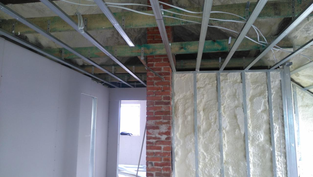 Wnętrze w trakcie prac wykończeniowych: ściana z płyt gipsowo-kartonowych, fragment ściany z cegły oraz ściana z metalową konstrukcją wypełnioną białą pianą izolacyjną. Widoczne elementy instalacji...