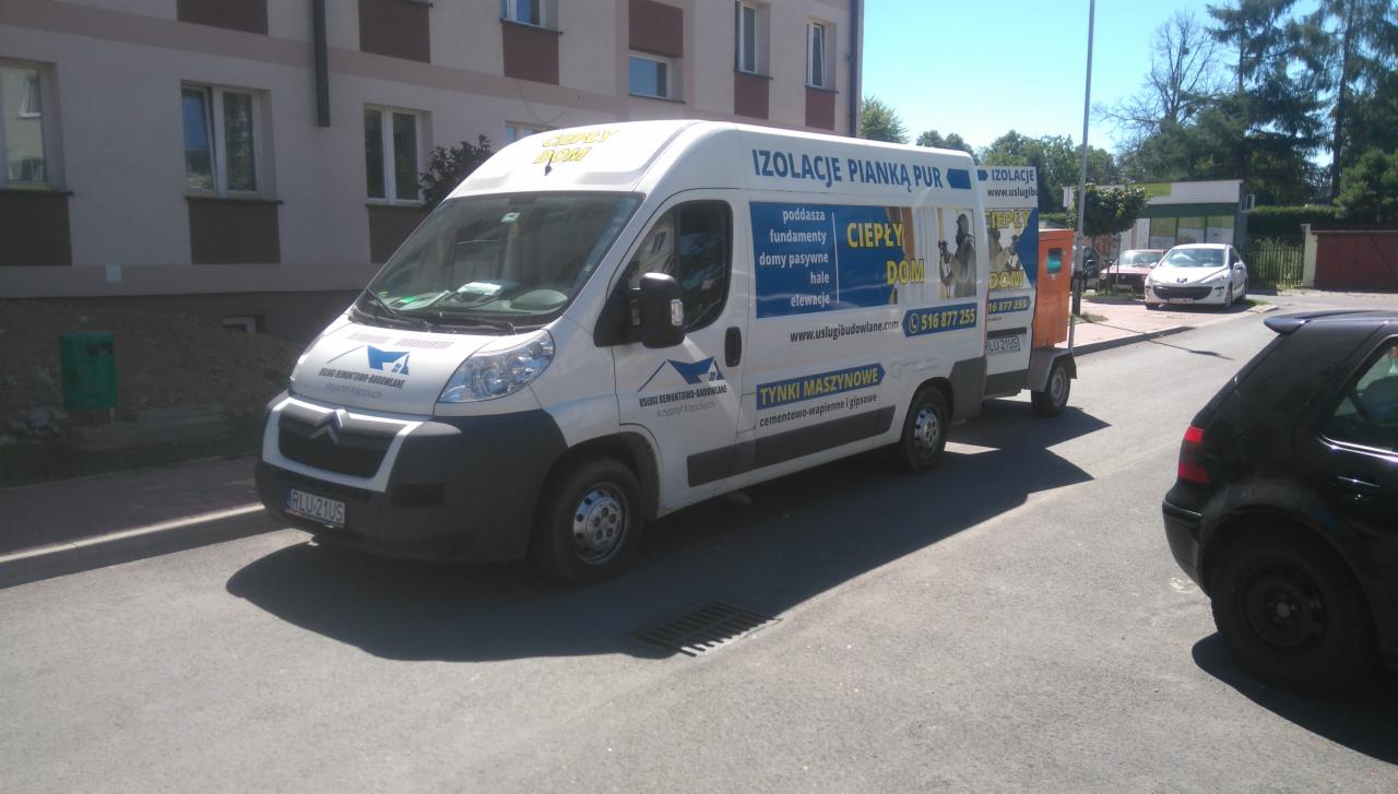 Biały samochód dostawczy z reklamą izolacji pianką PUR, zaparkowany na ulicy w słoneczny dzień, widoczne logo firmy i dane kontaktowe.