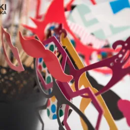 Kolorowe rekwizyty do fotobudki: wąsy, okulary i inne gadżety na patyku, umieszczone w czarnym, ażurowym pojemniku.