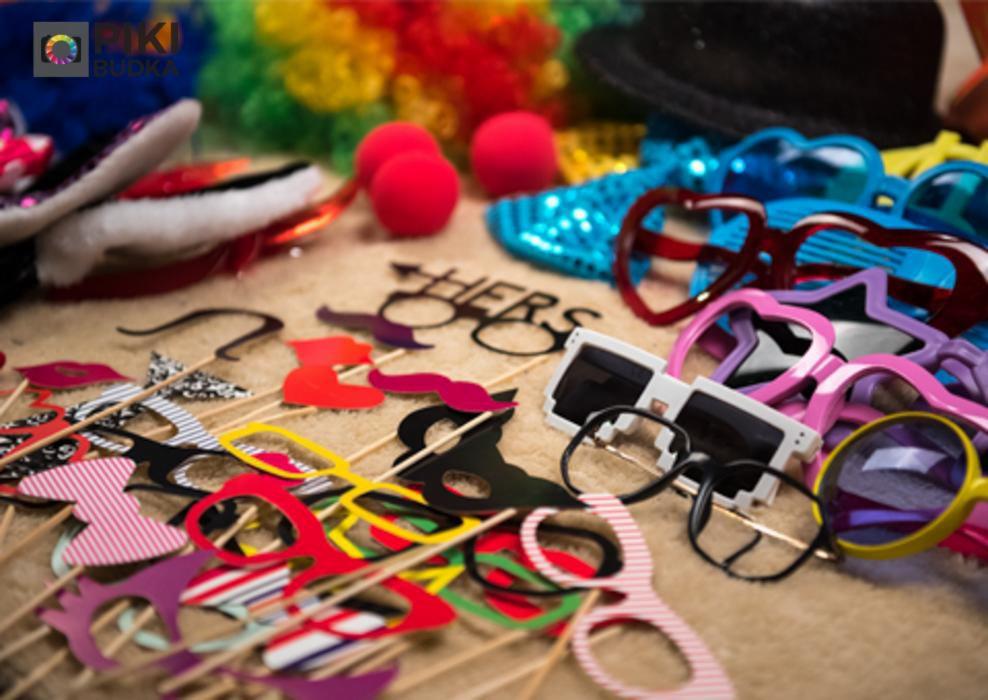 Różnorodność rekwizytów do fotobudki: kolorowe okulary, wąsy, usta i inne akcesoria na drewnianych patyczkach, kule klauna i kapelusz.