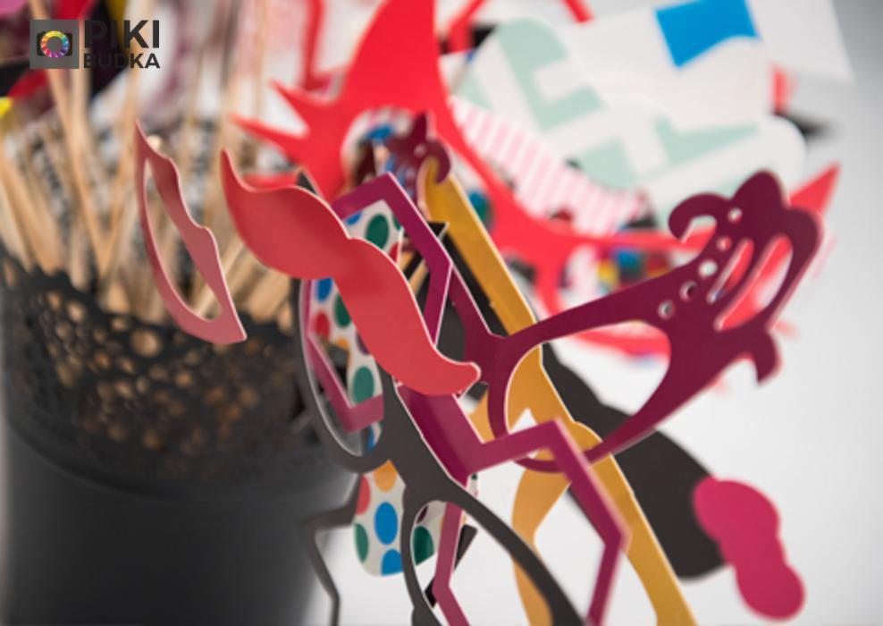 Kolorowe rekwizyty do fotobudki: wąsy, okulary i inne gadżety na patyku, umieszczone w czarnym, ażurowym pojemniku.