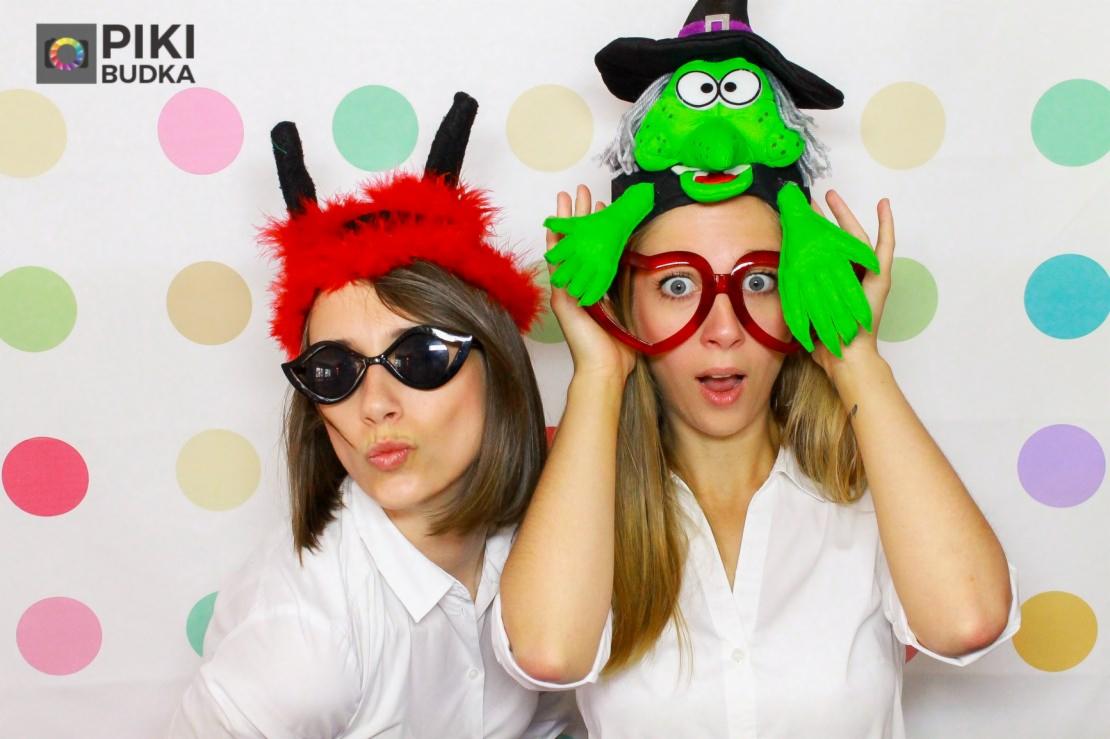 Dwie kobiety pozują w fotobudce z rekwizytami: czerwone rogi diabła i okulary przeciwsłoneczne oraz zielona czarownica na głowie i czerwone okulary, tło w kolorowe kropki.