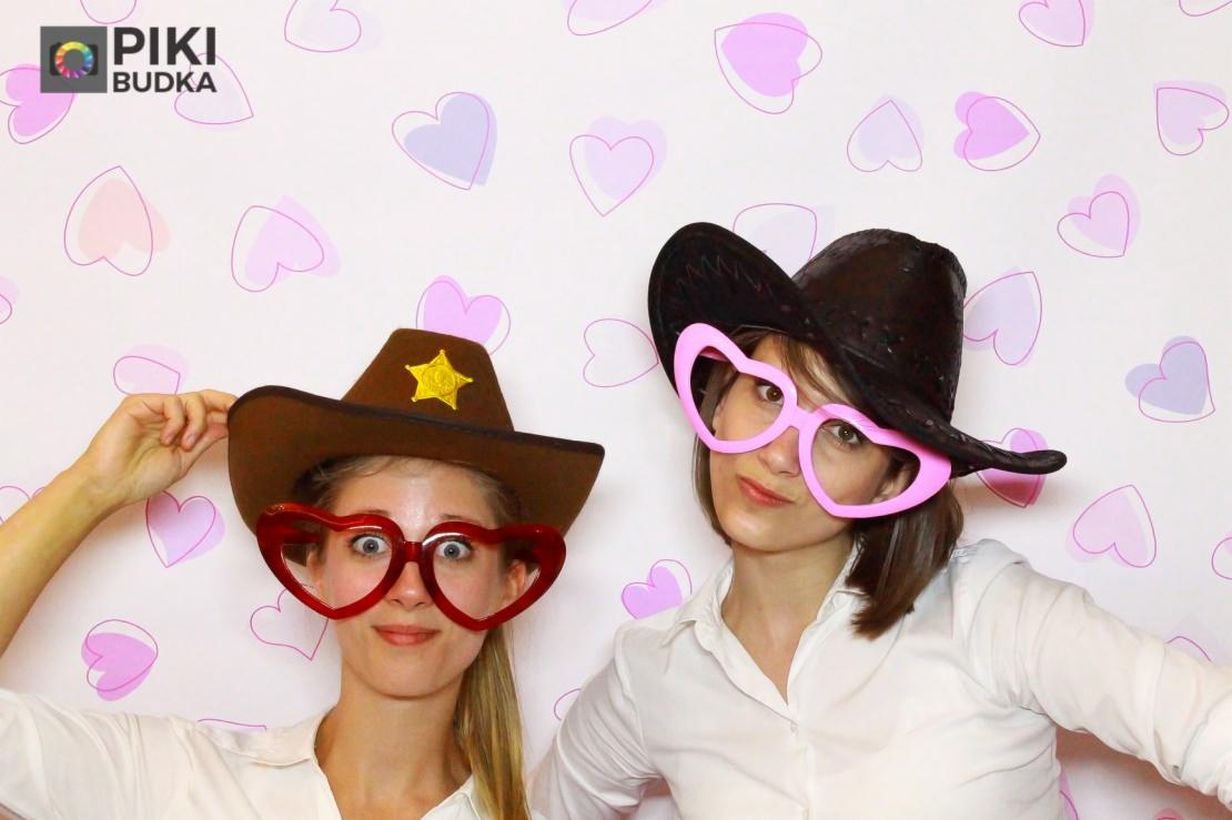 Dwie kobiety w fotobudce pozują w kapeluszach kowbojskich i okularach w kształcie serc na tle ściany z motywem serc.