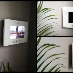 Biały wideofon Vidos zamontowany na szarej ścianie, widok z boku z fragmentem logotypu firmy Sofisto, dodatkowo ujęcie z bliska z elementami dekoracyjnymi z liści palmowych.