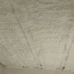 Poddasze pokryte grubą warstwą białej pianki izolacyjnej, widoczne elementy konstrukcyjne, nier&oacute;wnomierna struktura pianki.