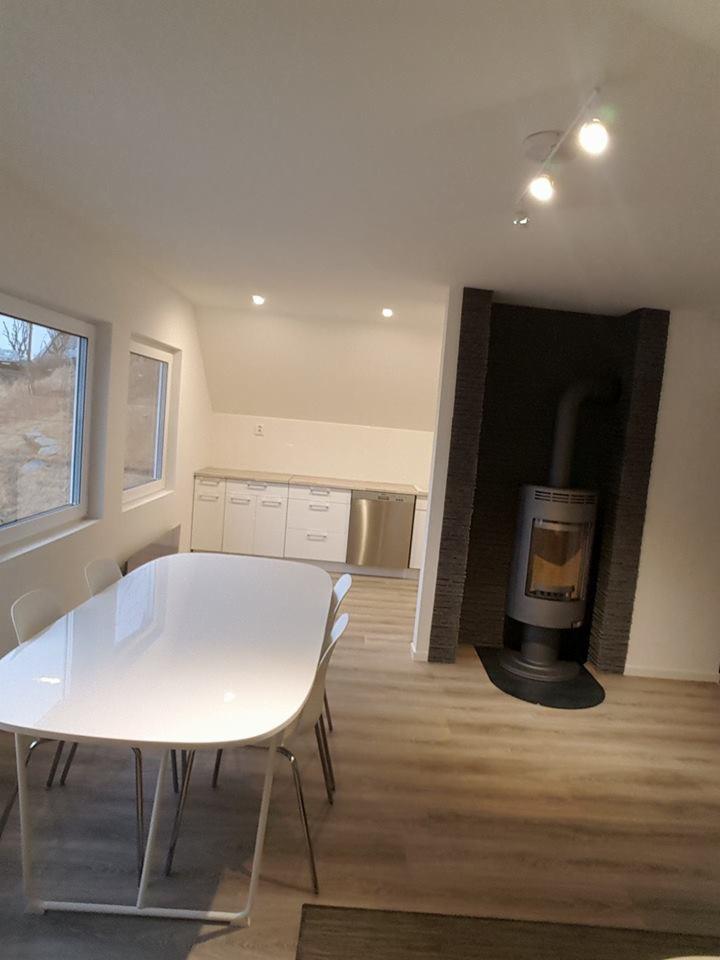 Jasne wnętrze z białym stołem jadalnym, piecem wolnostojącym obudowanym kamieniem dekoracyjnym i widokiem na zabudowę kuchenną w tle.