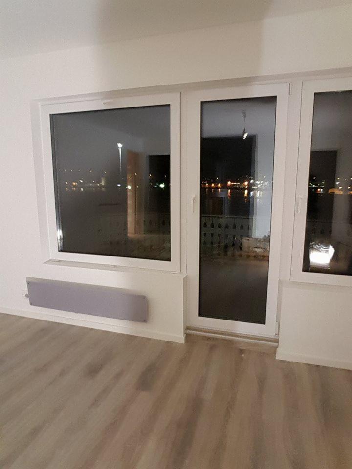 Wnętrze z nowymi oknami i drzwiami balkonowymi PCV, widok na nocne miasto, jasne panele podłogowe i szary grzejnik na ścianie.