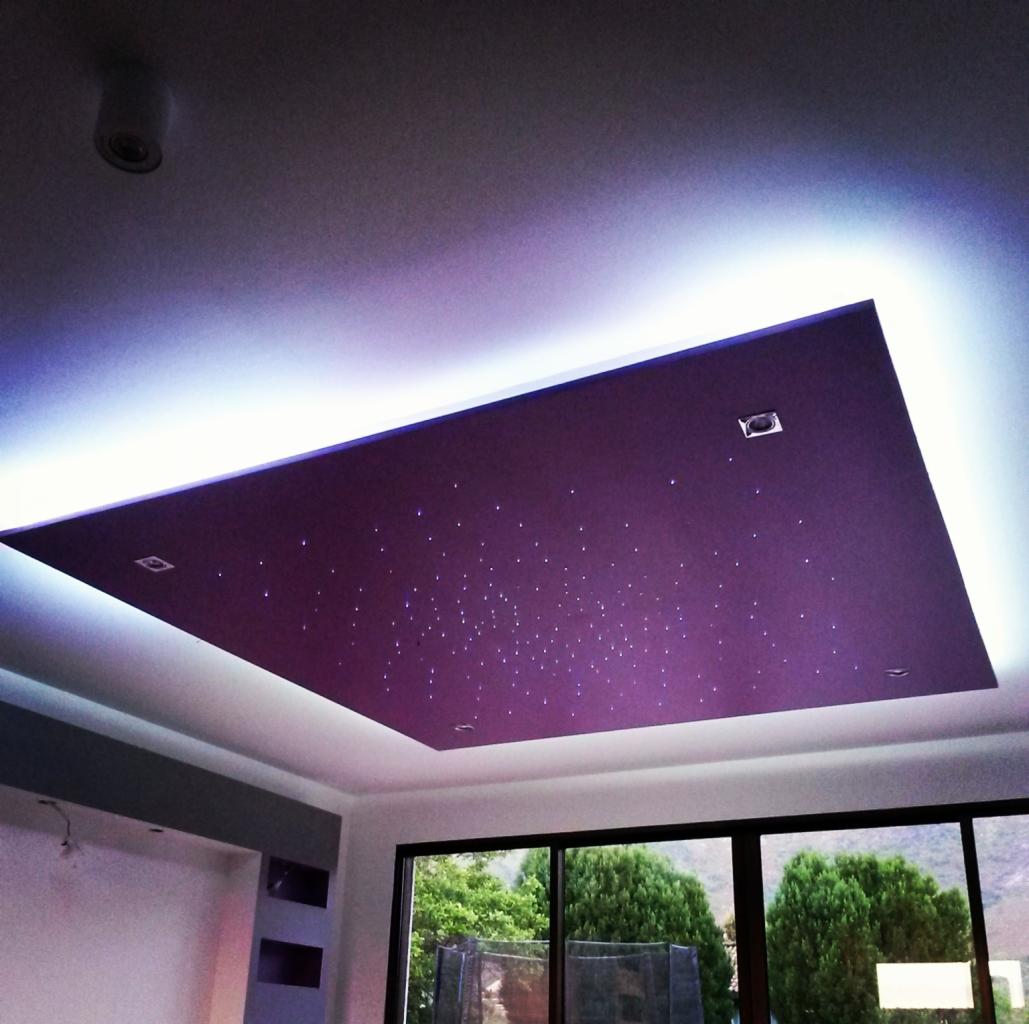 Fioletowy podwieszany sufit z efektem gwiazd, otoczony białym oświetleniem LED, widoczne cztery wbudowane reflektory punktowe, okno z widokiem na zieleń w tle.