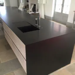 Długi, czarny blat kuchenny z wbudowanym zlewem i drewnianymi szafkami, na tle jasnego wnętrza z kamienną podłogą.