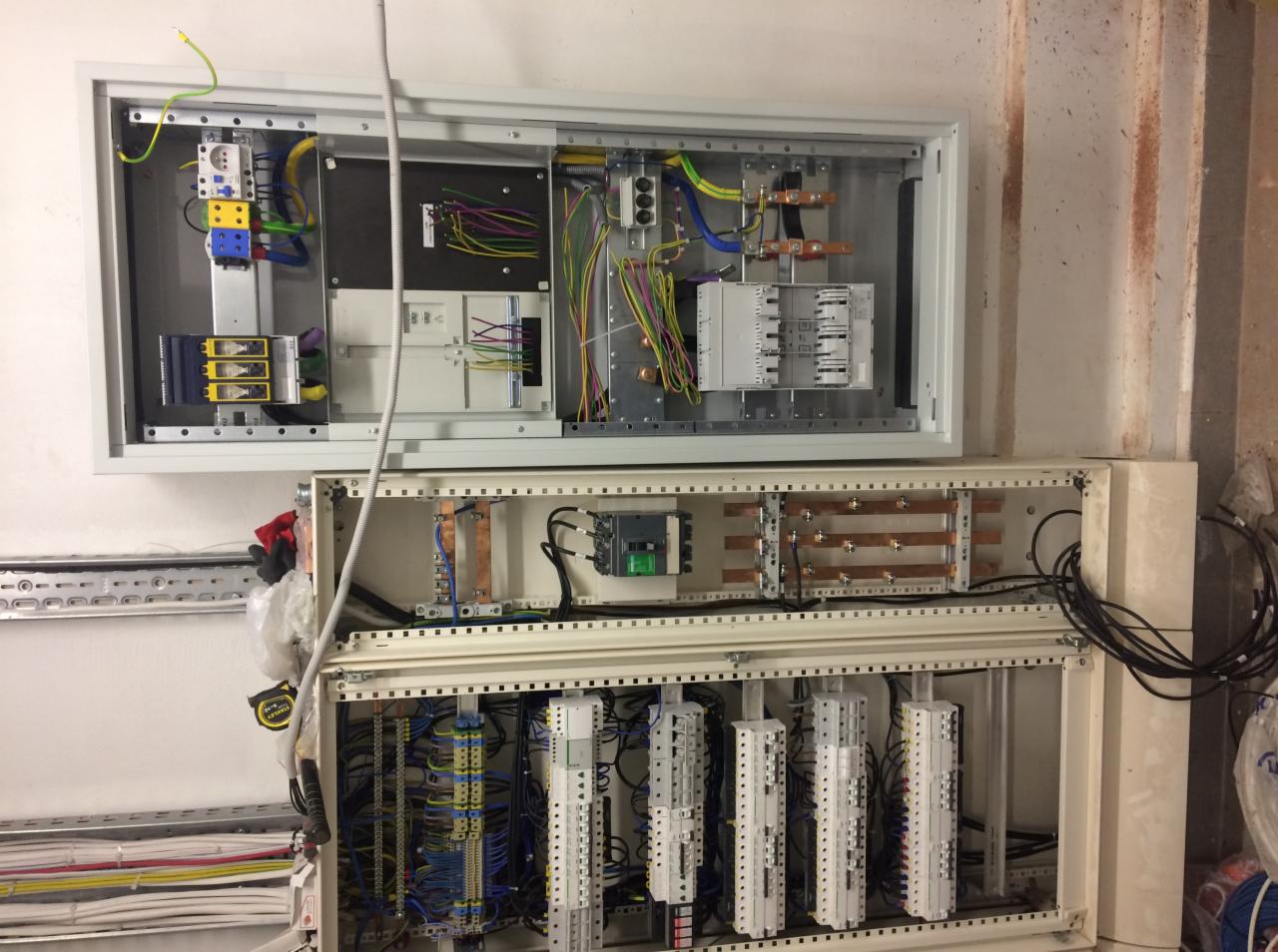 Dwie rozdzielnice elektryczne zamontowane na ścianie: górna z elementami w trakcie podłączania, dolna z uporządkowanymi przewodami i modułami, widoczne szyny miedziane i okablowanie wielokolorowe.