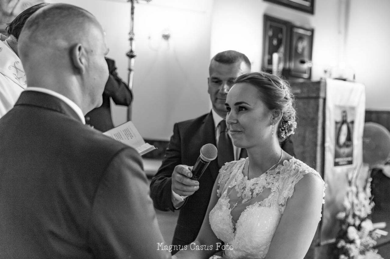 Panna młoda w koronkowej sukni słucha przysięgi małżeńskiej, mikrofon trzymany przez urzędnika, czarno-biała fotografia z kościelnego wnętrza.