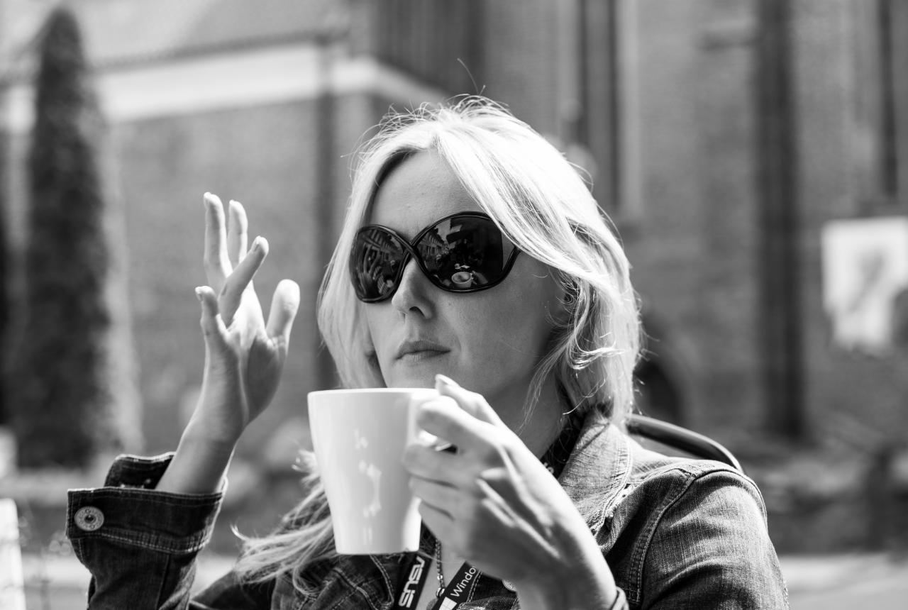Portret kobiety w okularach przeciwsłonecznych, trzymającej biały kubek, z odbiciem ulicy w szkle, w czarno-białej tonacji.
