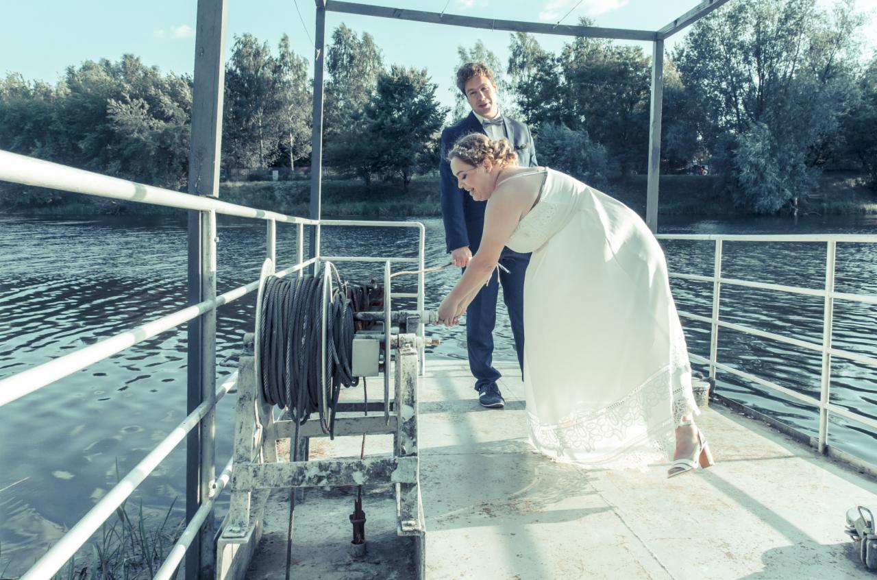 Para młoda na promie: Panna młoda w białej sukni i szpilkach próbuje uruchomić linę od promu, pan młody w granatowym garniturze stoi za nią, na tle rzeki i drzew.