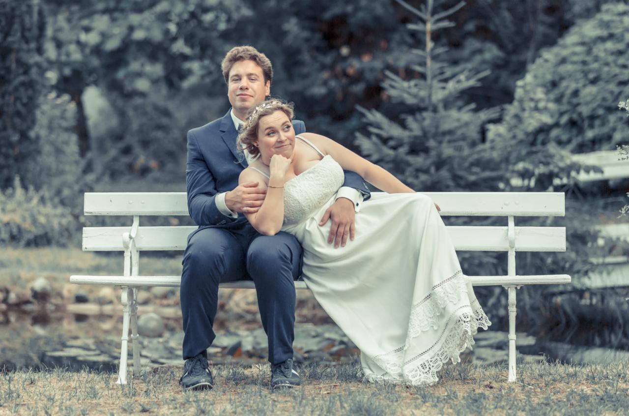 Para młoda siedzi na białej ławce w parku, panna młoda w koronkowej sukni spoczywa na kolanach pana młodego w granatowym garniturze, oboje patrzą w bok.
