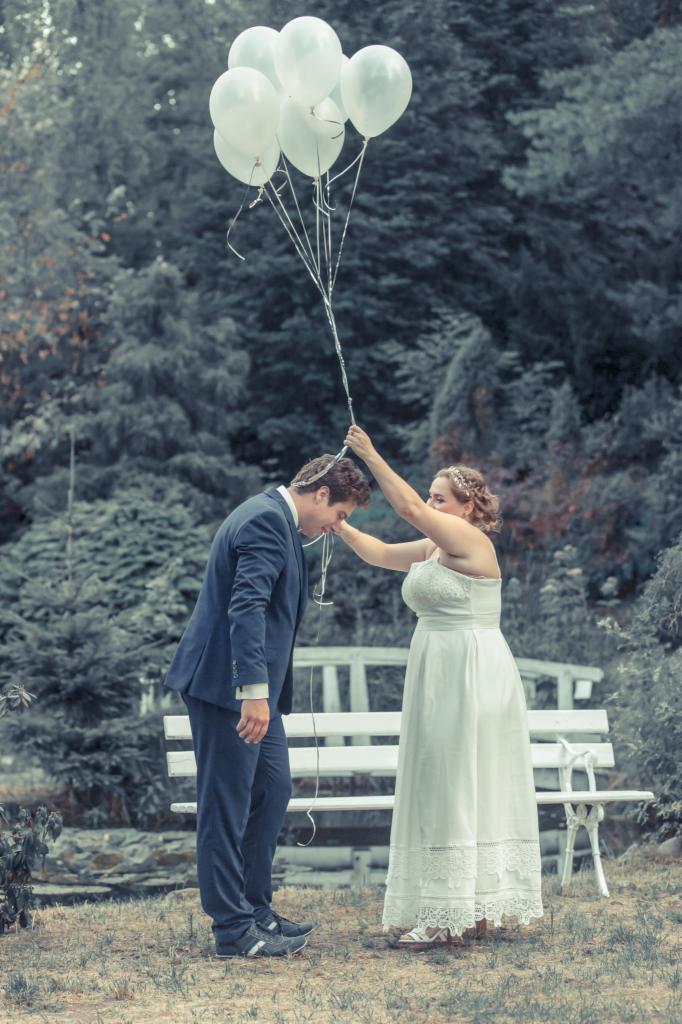 Panna młoda trzyma bukiet białych balonów nad głową pochylonego pana młodego w granatowym garniturze, na tle parku z białą ławką.