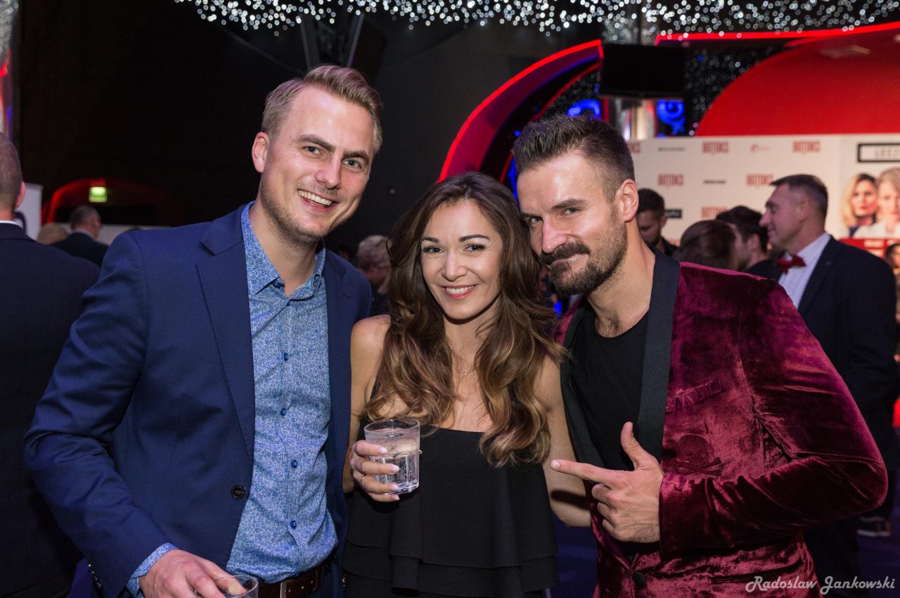 Trzy osoby uśmiechnięte pozują na tle czerwono-czarnej dekoracji na wydarzeniu, mężczyzna w bordowej marynarce wskazuje palcem w bok.