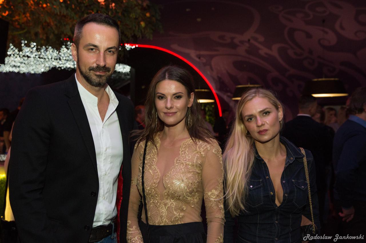 Grupa trzech osób pozuje na tle ciemnego wnętrza z dekoracyjnym oświetleniem i wzorzystą ścianą.