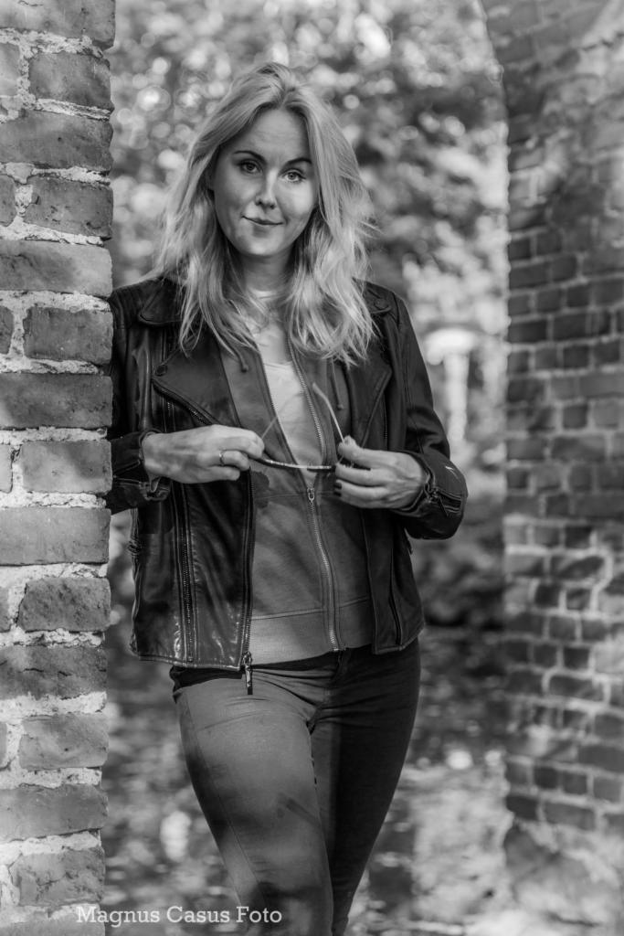 Portret kobiety w skórzanej kurtce, opierającej się o ceglany mur z okularami w dłoni, na tle rozmytej zieleni.