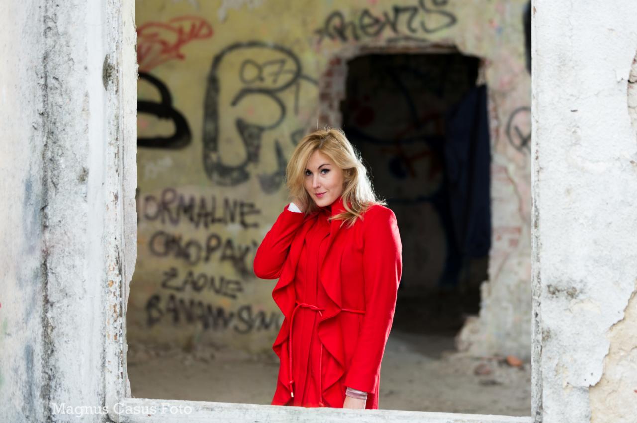Portret blondwłosej kobiety w czerwonym płaszczu, pozującej w zrujnowanym budynku z graffiti, ujęcie zza betonowej ramy okiennej.