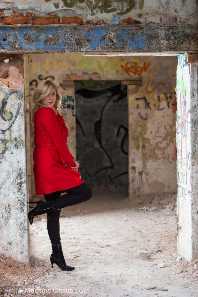 Kobieta w czerwonym płaszczu i czarnych legginsach pozuje w opuszczonym budynku z graffiti na ścianach.