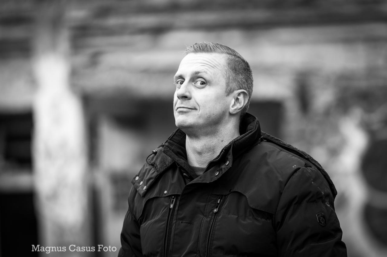 Portret mężczyzny w czarnej kurtce na rozmytym, miejskim tle, wykonany w czerni i bieli, z charakterystycznym spojrzeniem w bok.