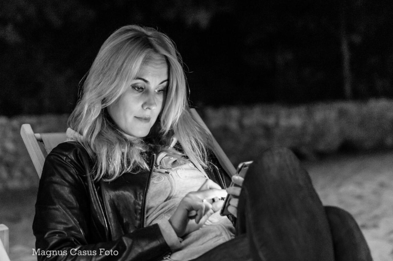 Czarno-białe zdjęcie kobiety siedzącej na leżaku, spoglądającej na telefon komórkowy, z widocznym logo fotografa w lewym dolnym rogu.