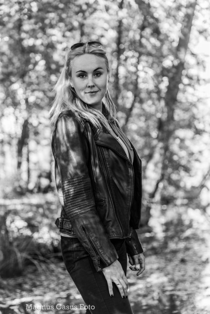 Portret kobiety w skórzanej kurtce na tle zamazanego lasu, czarno-białe zdjęcie z widocznym logo fotografa w rogu.