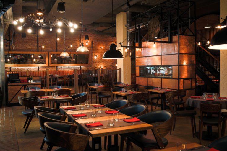 Wnętrze restauracji Salina z drewnianymi stołami nakrytymi czerwonymi serwetkami, oświetlone industrialnymi lampami i z dekoracyjną ścianą z miedzi.