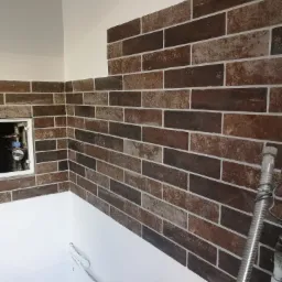 Brązowa ściana wyłożona kafelkami imitującymi cegłę, biała fuga, wnęka z wodomierzem, częściowo pomalowana ściana, instalacja hydrauliczna.