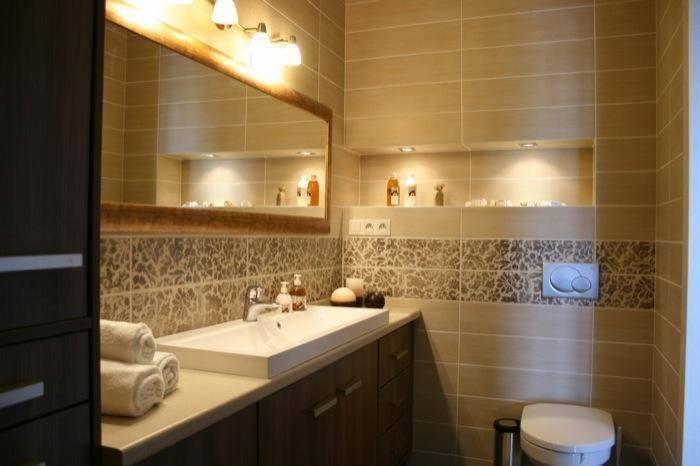 Wykończona łazienka w beżowych tonacjach z dekoracyjnymi płytkami, prostokątną umywalką nablatową, dużym lustrem z oświetleniem i podświetlaną wnęką na kosmetyki.